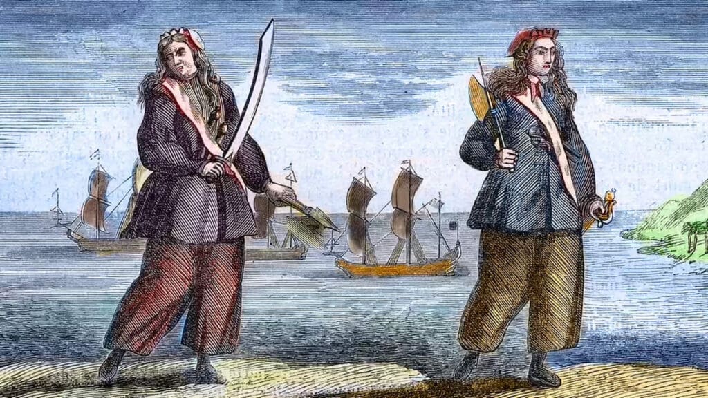 Illustrated depiction of Anne Bonny and Mary Read, two 18th-century female pirates, standing on a shoreline with swords in hand. Both are dressed in loose trousers and pirate-style coats, with sailing ships visible in the background under a cloudy sky.